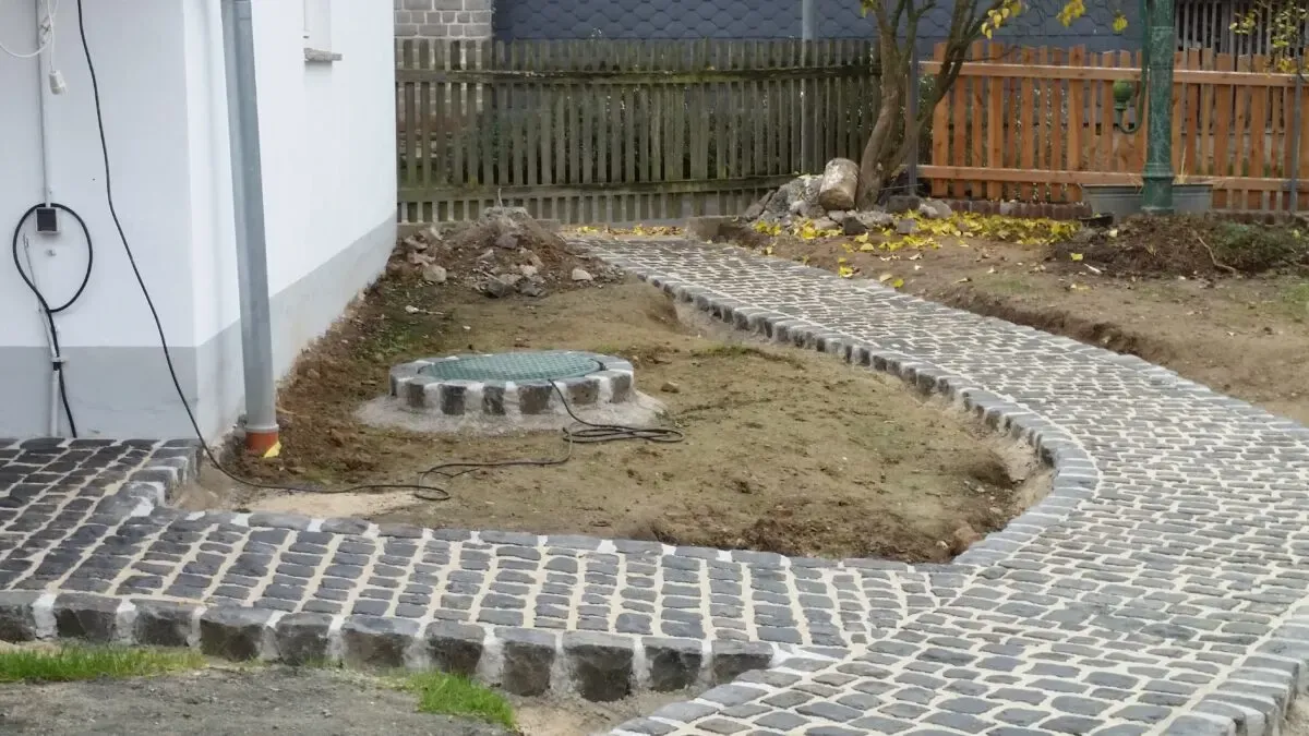 Geschwungener Gartenweg aus Granit-Kleinpflaster mit Zisternendeckel im Beet