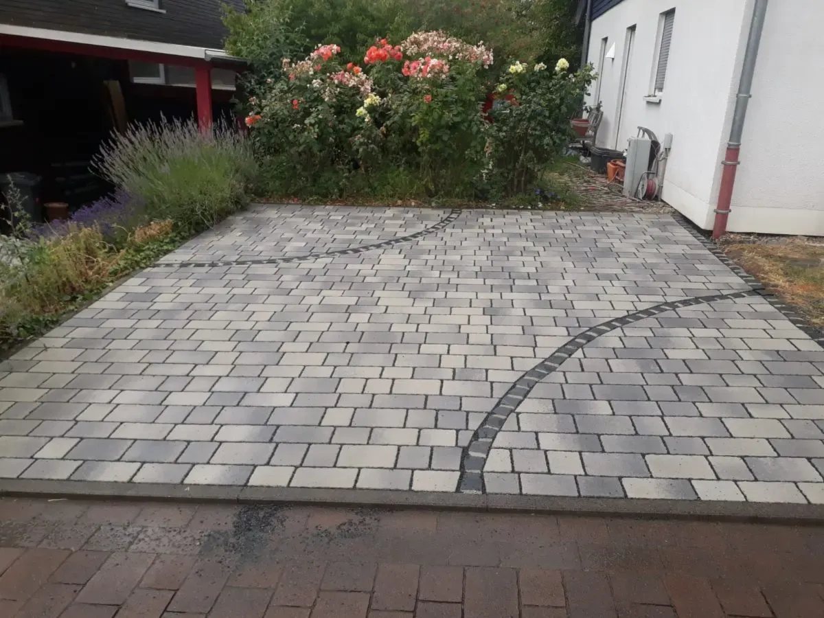 Hoffläche aus grauem Betonpflaster mit eingelegtem Bogen aus dunklem Kleinpflaster vor einem Rosenbeet